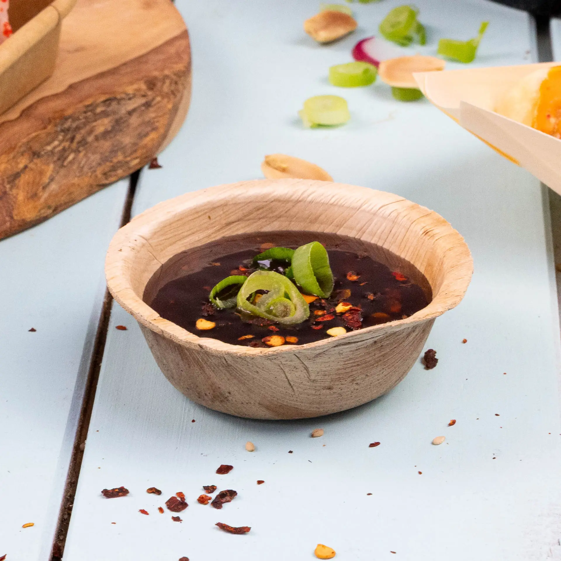 Round palm leaf bowl filled with dark sauce, topped with green onion slices and red pepper flakes, placed on a blue surface.