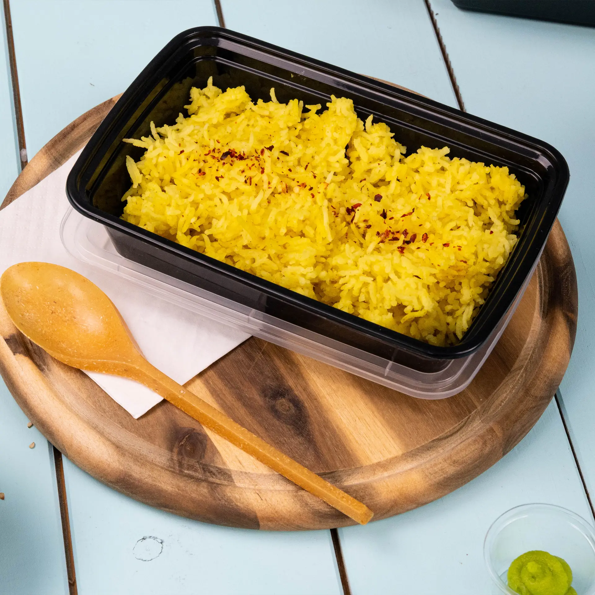 Reusable black rectangular container with yellow rice inside, wooden spoon beside, on wooden plate with white napkin.