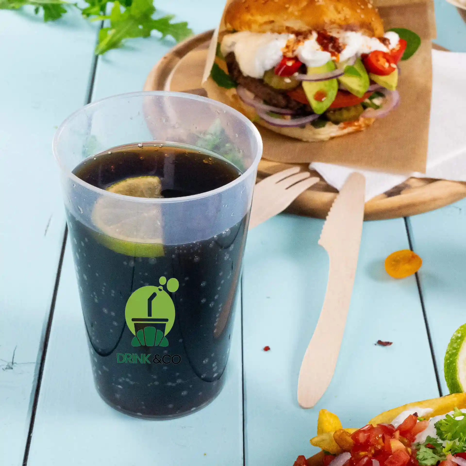 Clear reusable cup with logo, 500 ml capacity, filled with dark beverage and a lime slice, placed beside a burger and salad.