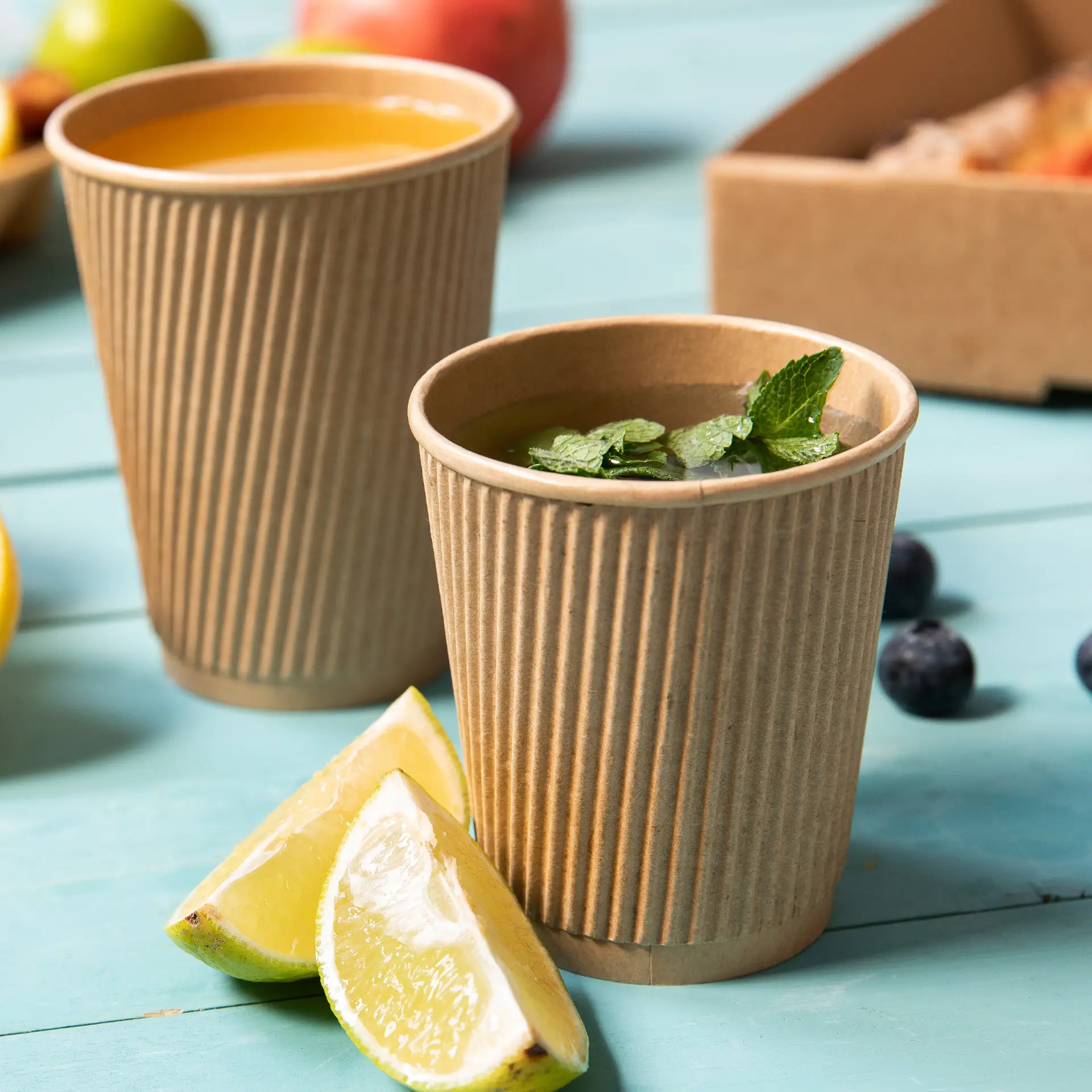 Two brown, ribbed disposable coffee cups on a blue table, one filled with tea and mint leaves, and the other with juice, beside lemon wedges.