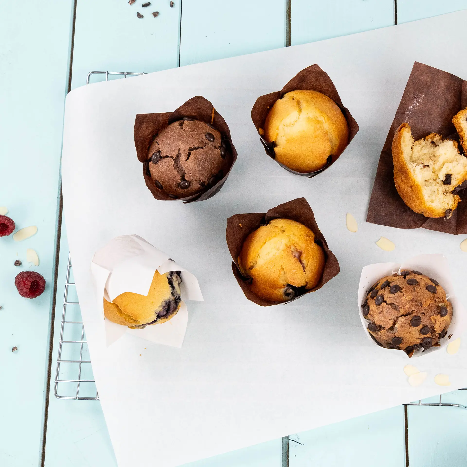 Assorted muffins in greaseproof brown paper cups on a white baking sheet, with some raspberries and almond slivers nearby.
