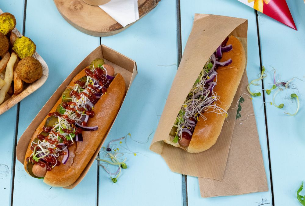 Two hot dogs with various toppings, including pickles, onions, and sprouts, served in paper containers on a blue table.