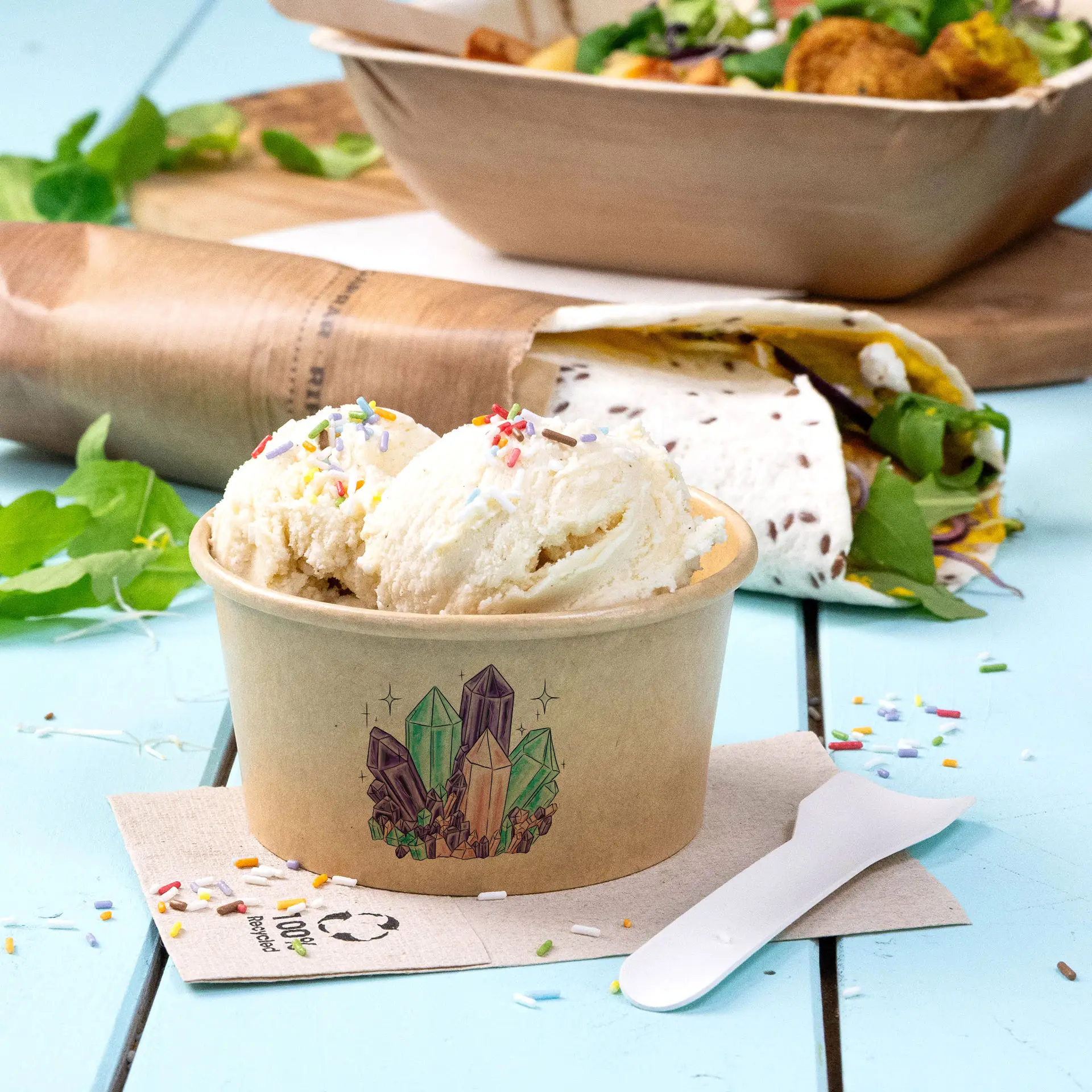 Ice cream in a brown printed cup with colorful sprinkles, placed on a blue surface with a napkin and greenery.