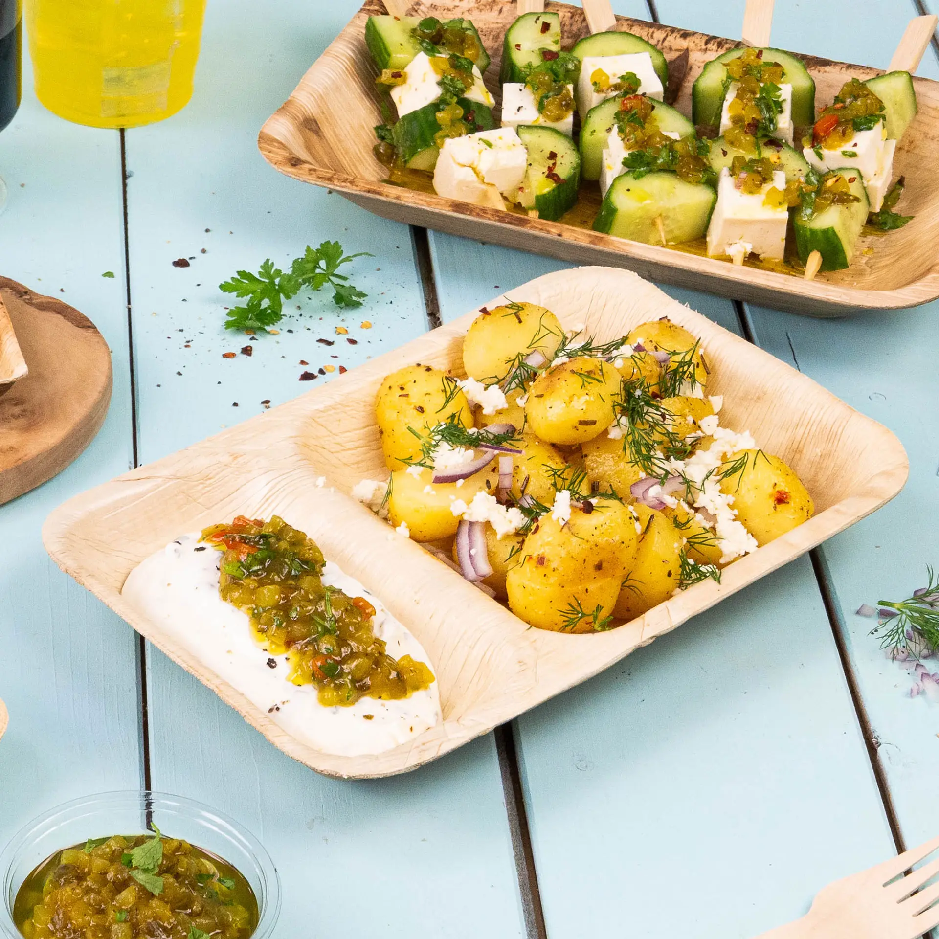 Three-compartment palm leaf menu plate with food items including hummus, sautéed vegetables, and dumplings on a light blue background.