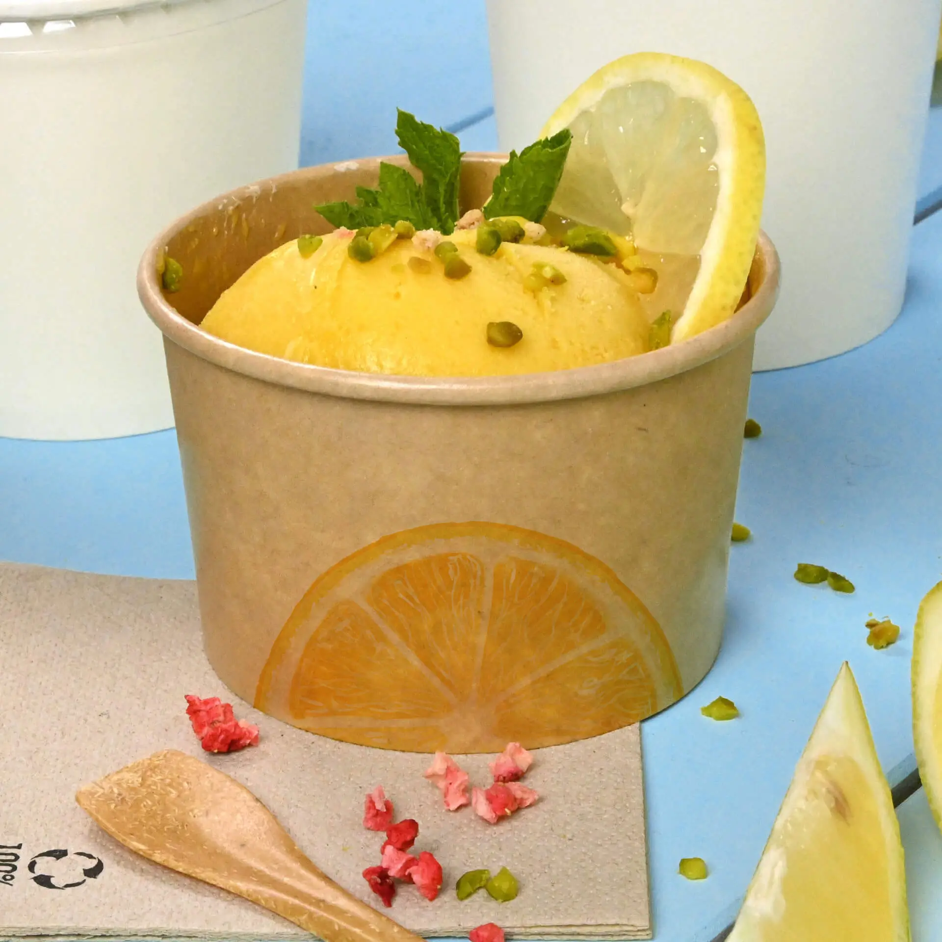 Brown ice cream cup with a lemon-flavored scoop, garnished with mint and pistachios, on a blue surface.
