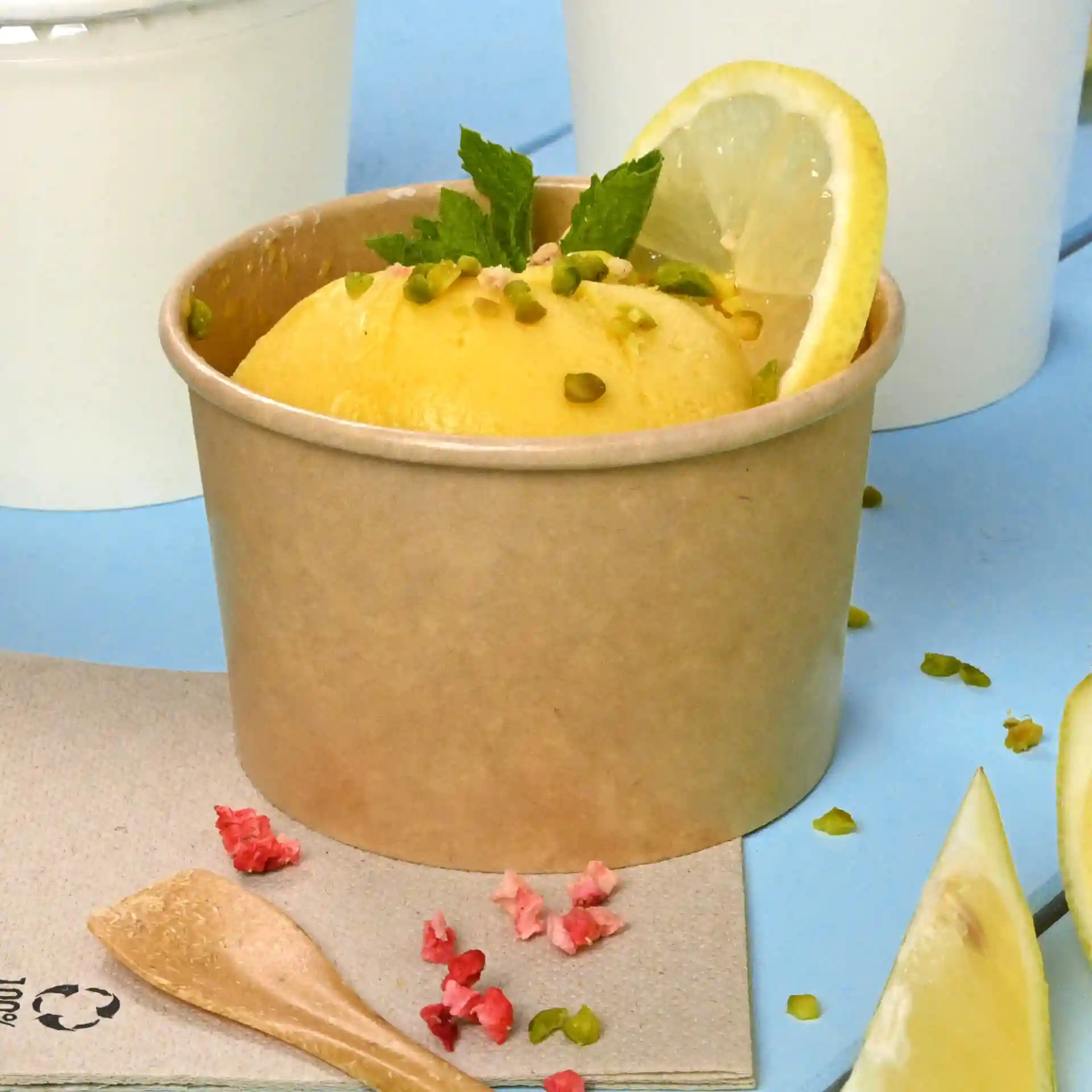 Brown paper ice cream cup filled with yellow ice cream, topped with lemon slice, mint, and pistachios, on a blue surface.