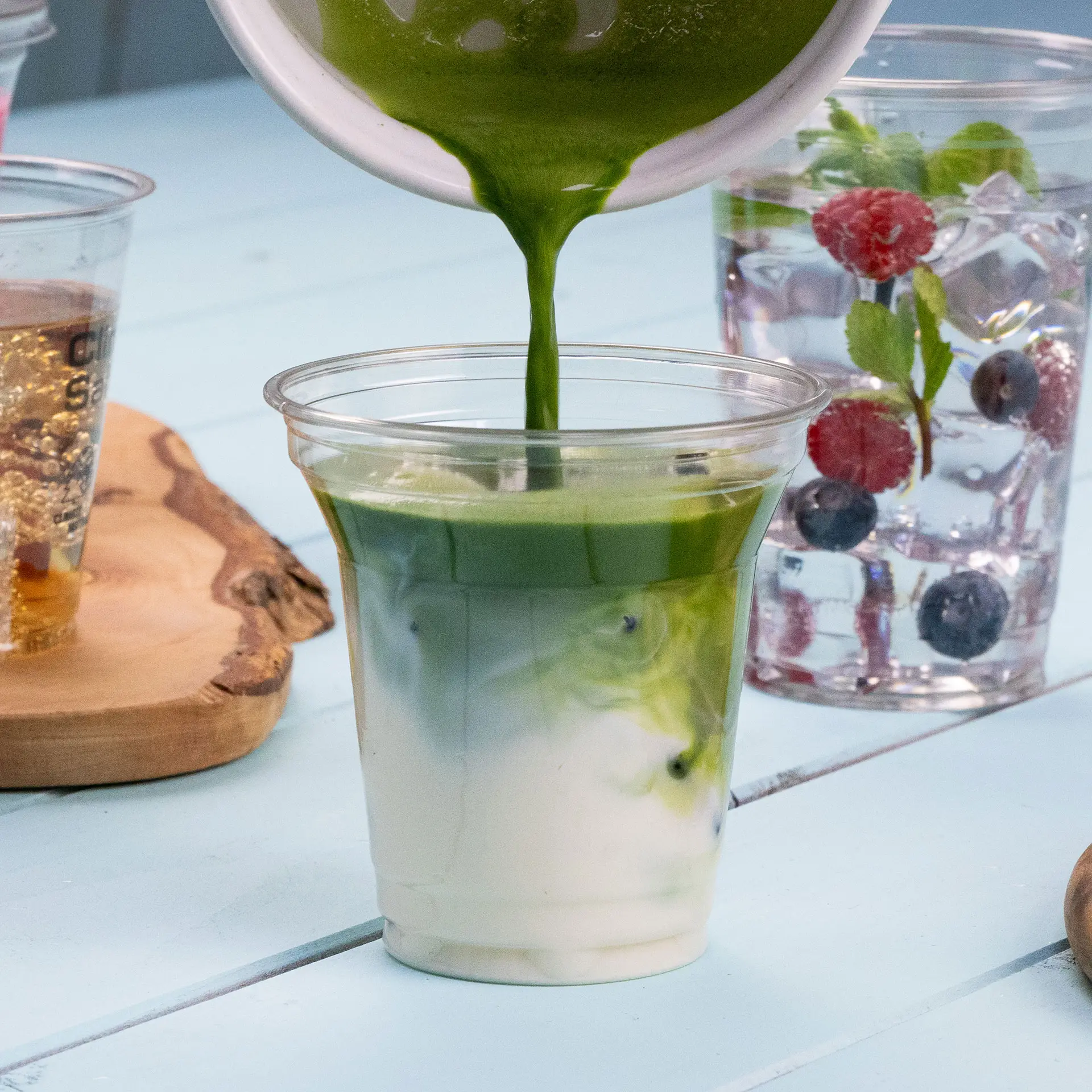 A clear rPET cup is being filled with a green beverage, showcasing layers of color; additional drinks are visible in the background.