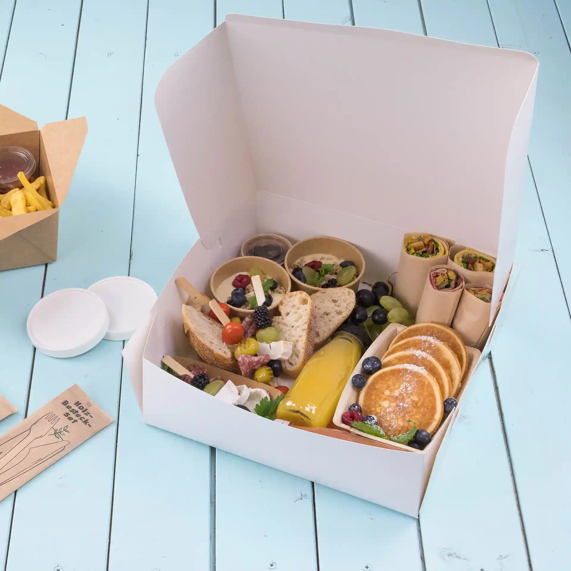 White cake box filled with assorted food items including bread, fruit, pancakes, and sauces, on a blue wooden surface.