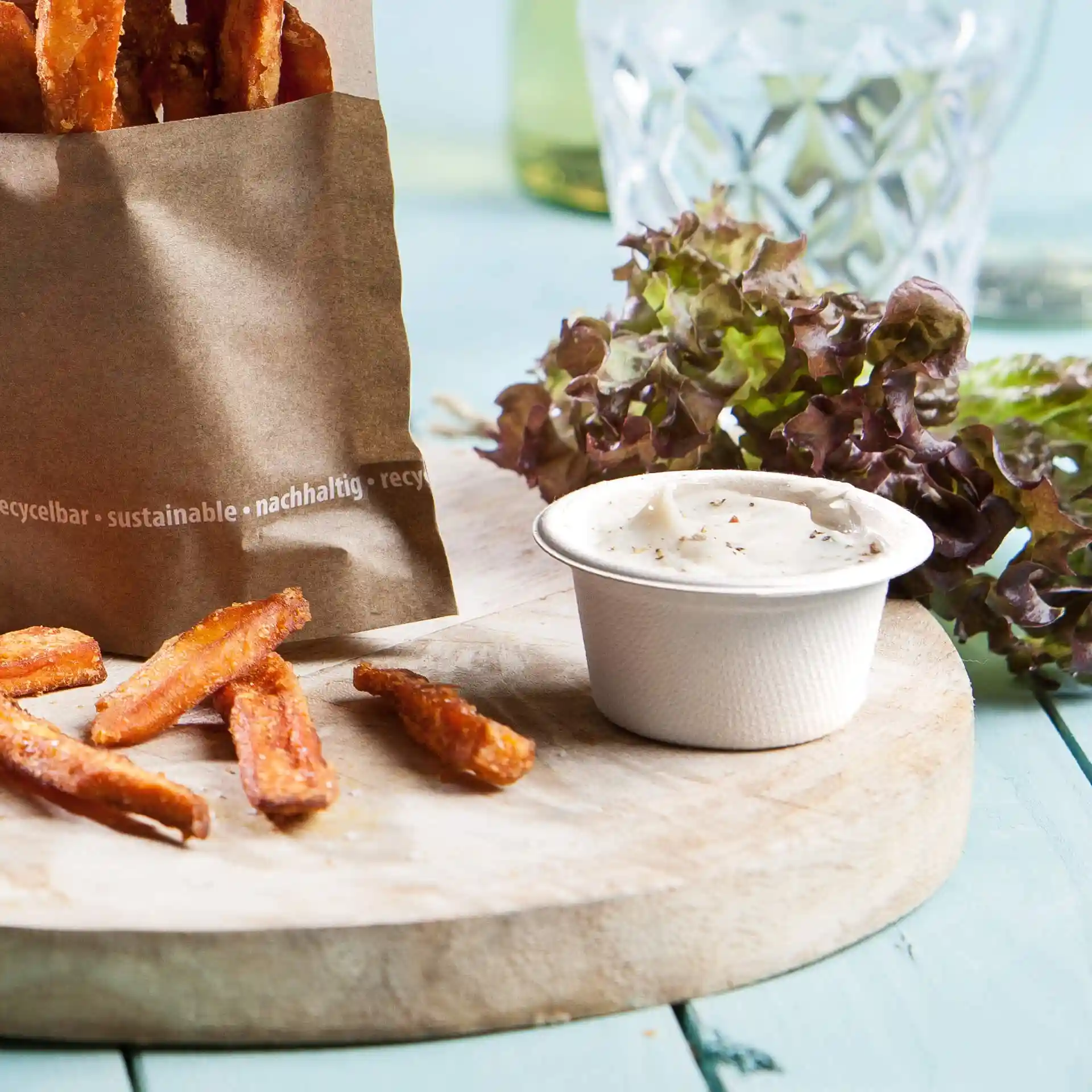Image of a paper bag with sweet potato fries, a small round cup of dip, and lettuce on a wooden board.