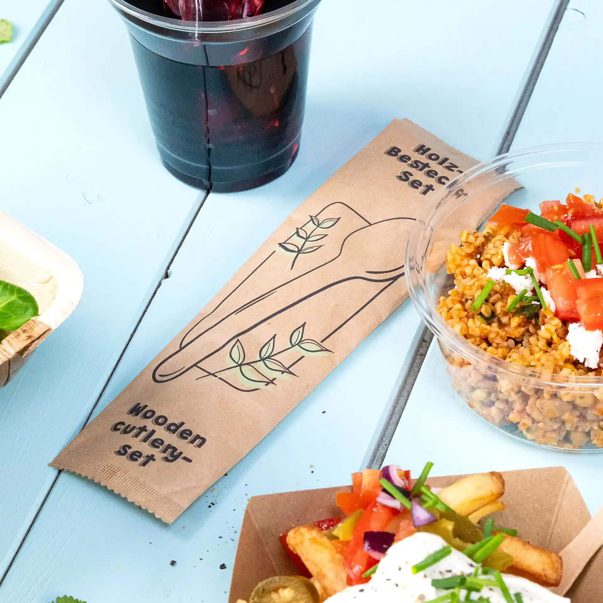 Wooden cutlery set packaging with a design of utensils and leaves, placed beside food items on a blue surface.