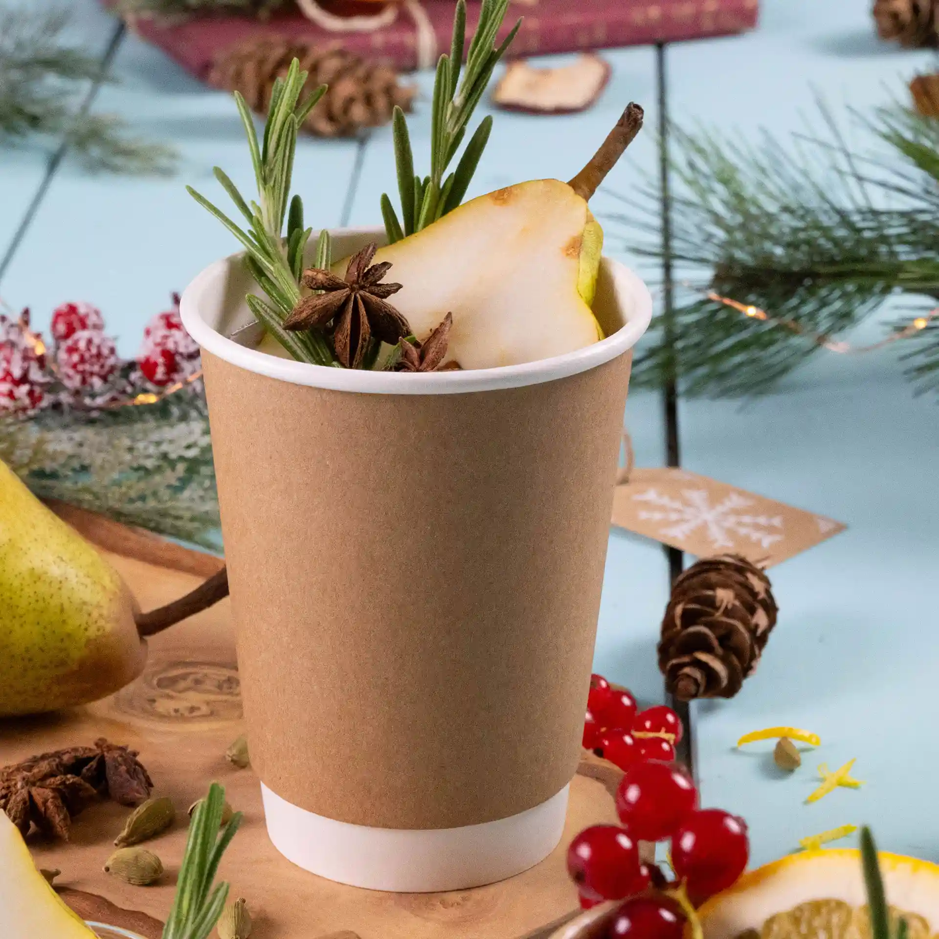 Brown double-walled cardboard cup filled with a pear slice, rosemary, and star anise, placed on a light blue surface.