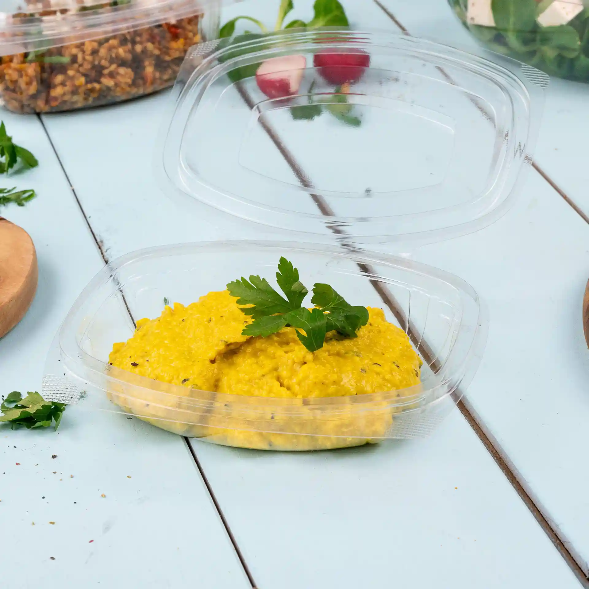 Transparent rectangular deli bowl with a folding lid, containing yellow food topped with a sprig of parsley.