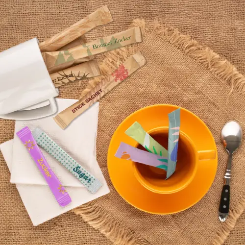 Colorful sugar stick sachets in a cup of tea on a rustic table setting with a spoon and empty cups.