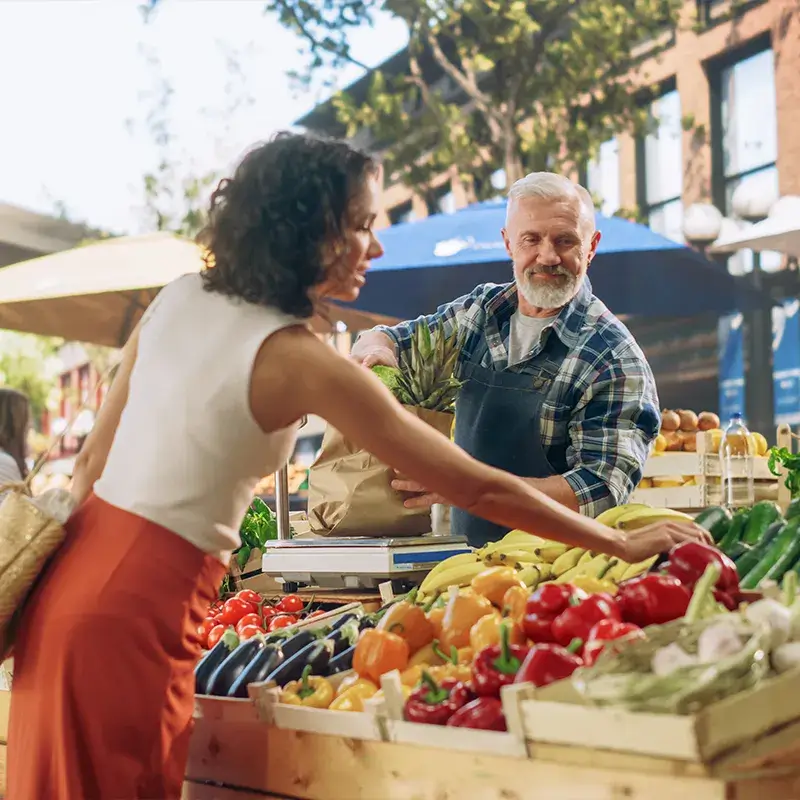 A woman in a red skirt purchases vegetables from a vendor at a market stall filled with fresh produce.