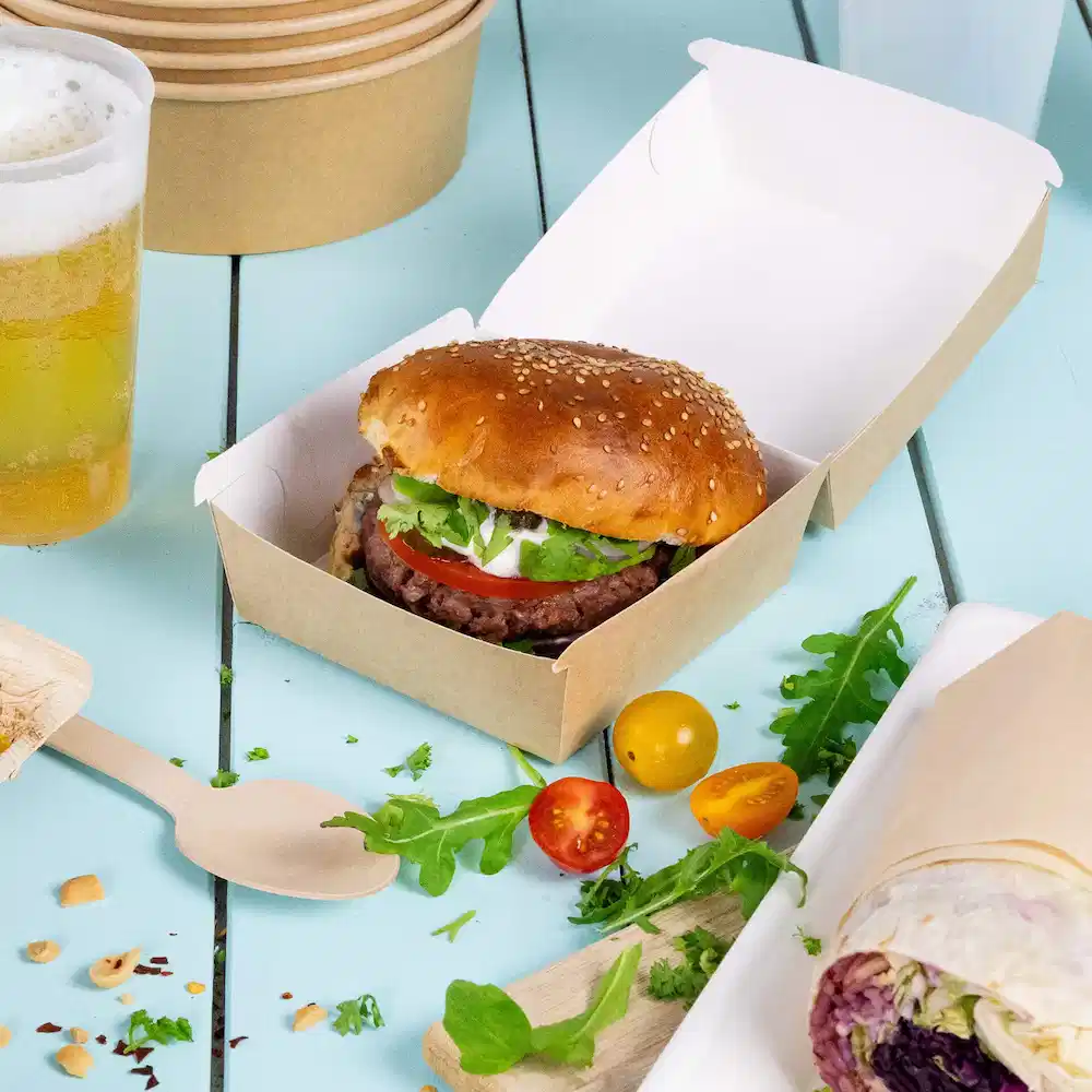 A sesame seed bun burger with lettuce, tomato, and sauce in a brown takeout box, accompanied by vegetables and beer.