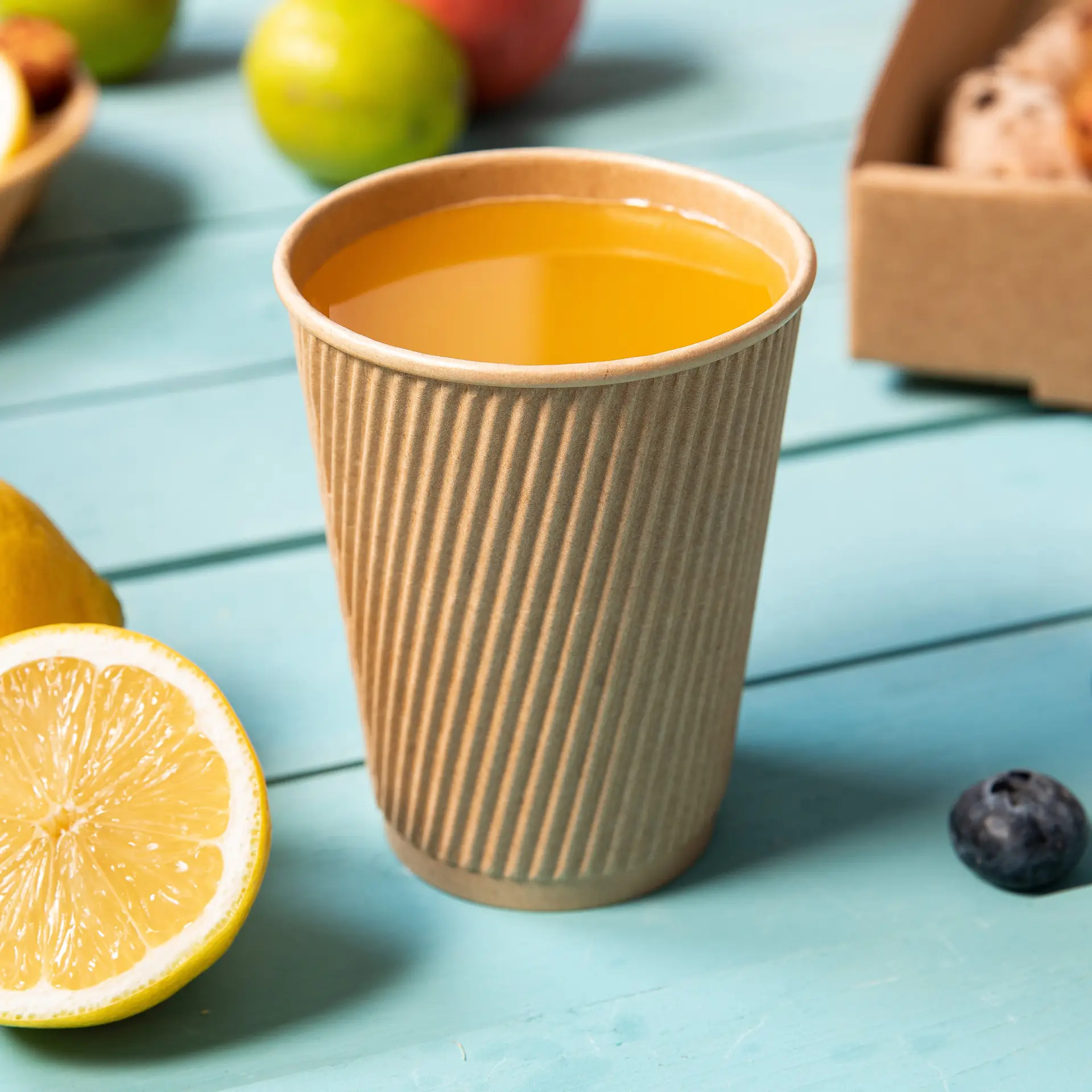 One brown ribbed disposable coffee cup with hot drink; lemon slice and blueberries nearby on a blue wooden table.