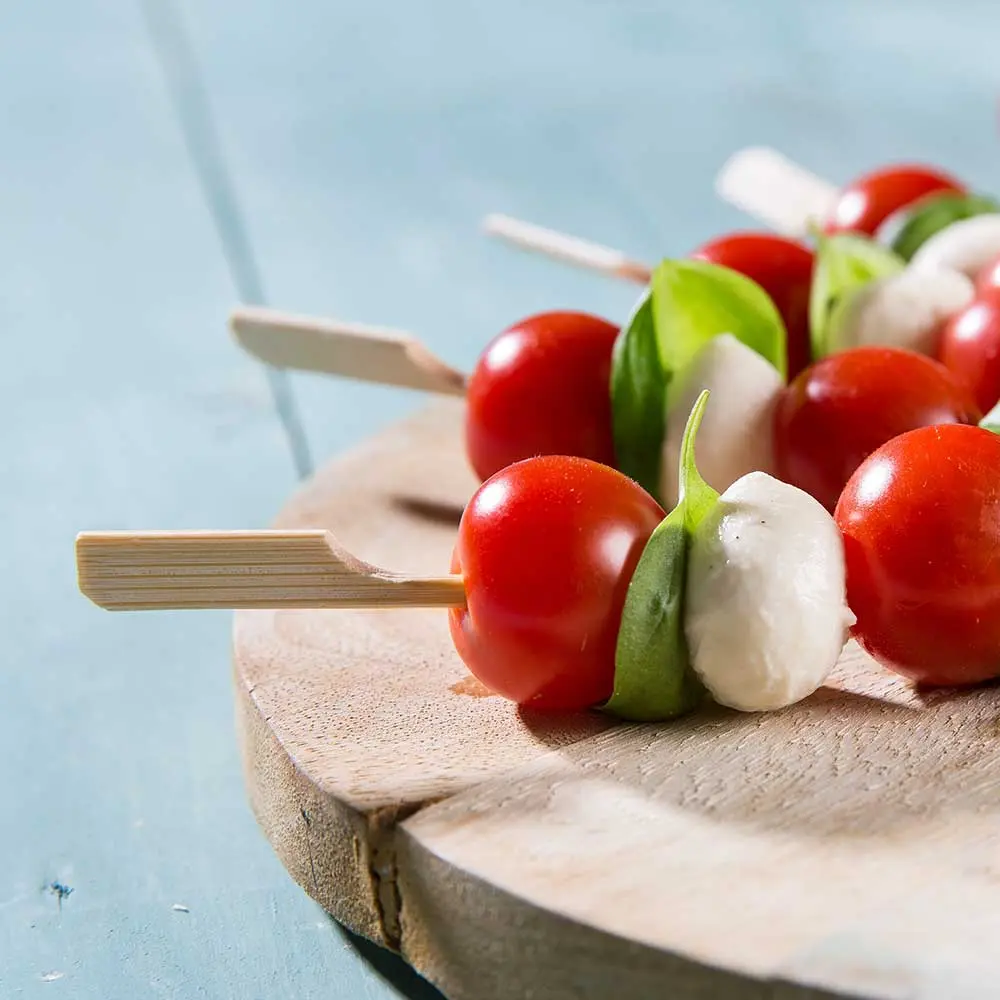 Skewers with cherry tomatoes, basil leaves, and mozzarella balls on a wooden board.