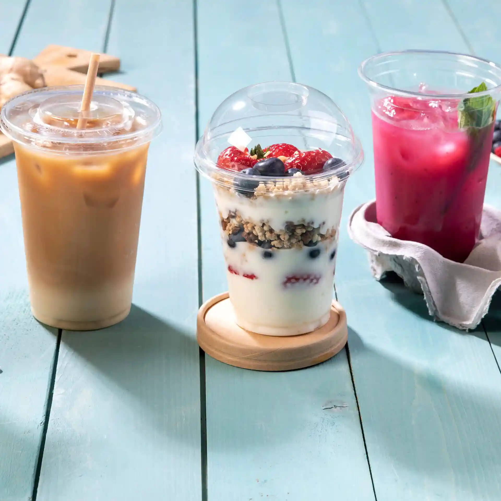 Image of three beverages: an iced coffee, a yogurt parfait with berries, and a pink drink, all in transparent cups on a blue wooden surface.