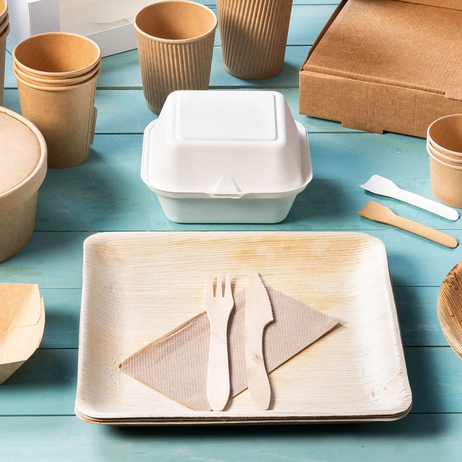 Image of a white takeout container, wooden utensils on a plate, and various paper cups and boxes on a blue surface.