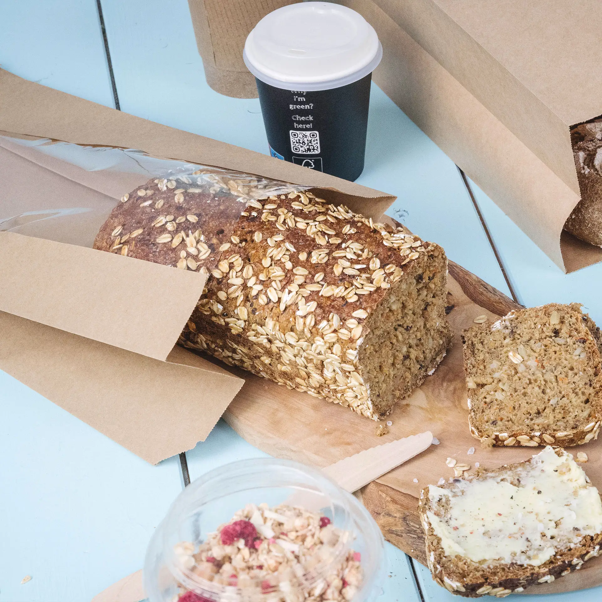 One loaf of brown bread with oatmeal topping, partially sliced, next to a coffee cup and a container of granola, all on a blue surface.
