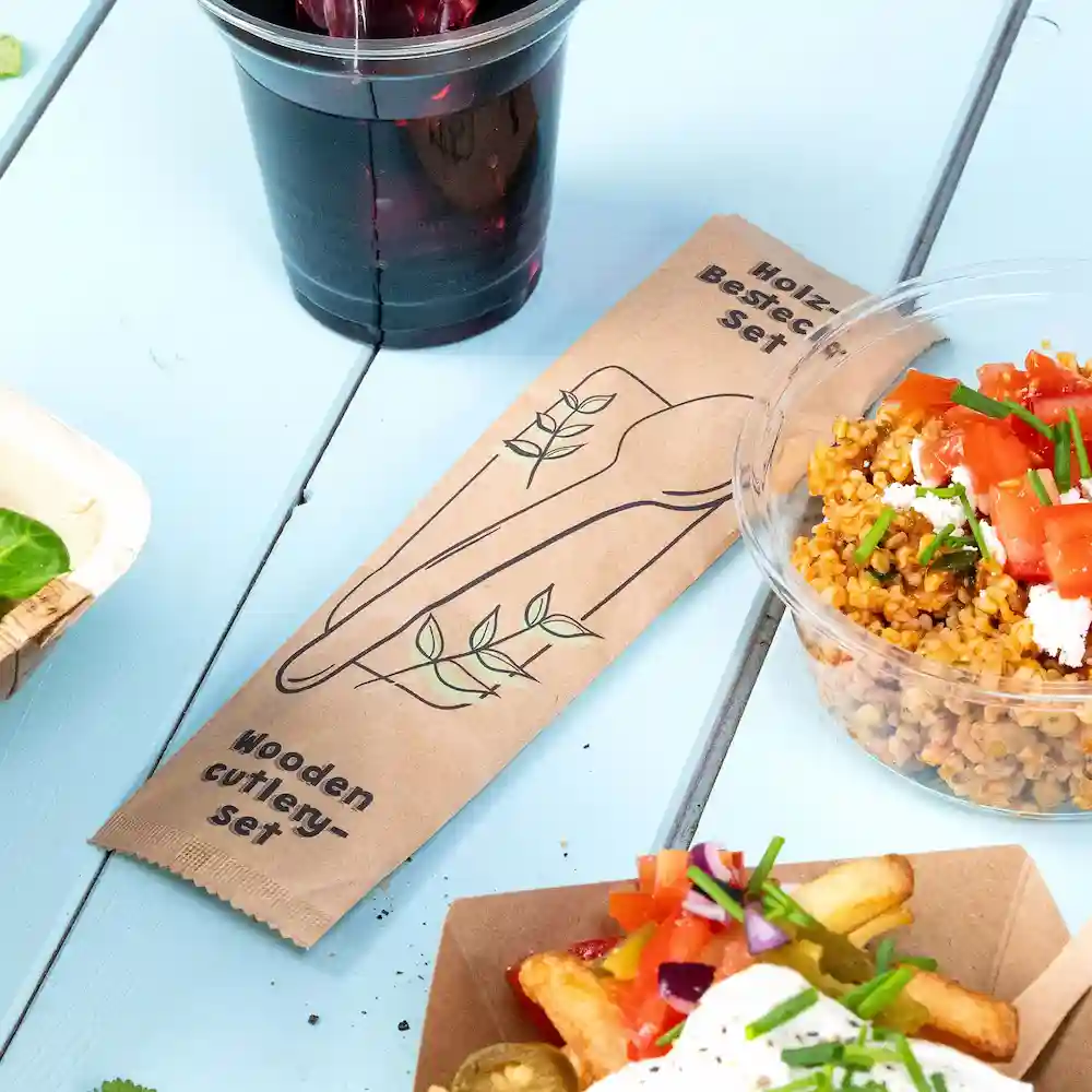 Wooden cutlery set packaging next to a plastic cup, a bowl of grain salad, and a cardboard container of fries on a blue surface.