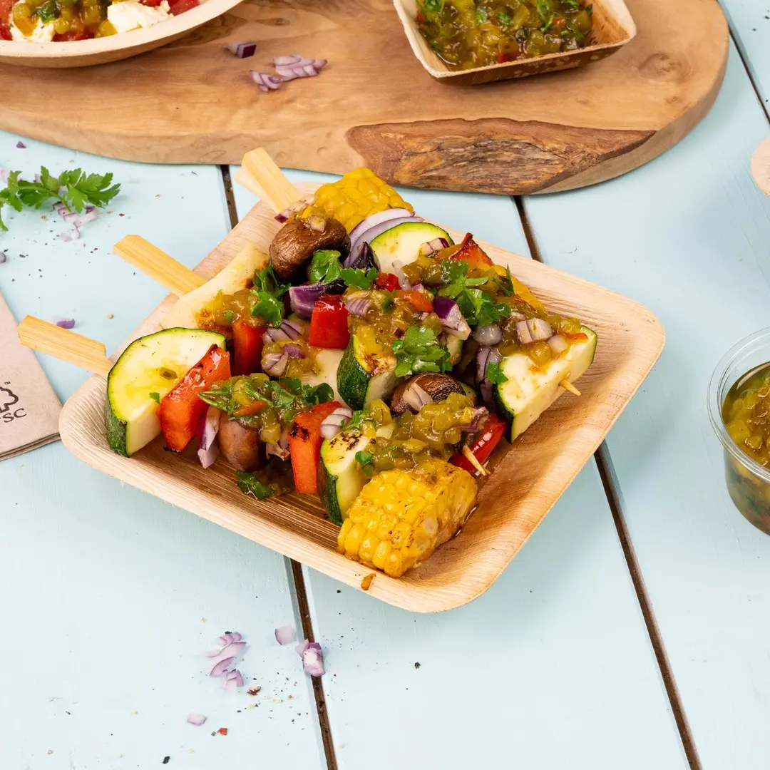 Wooden skewers with grilled vegetables including zucchini, bell pepper, corn, and mushrooms topped with sauce in a wooden tray.