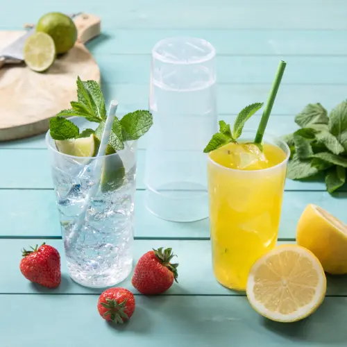Two reusable cups with drinks, one clear with a straw containing a lime and mint, and one yellow drink with a straw, surrounded by fresh strawberries and lemon slices.