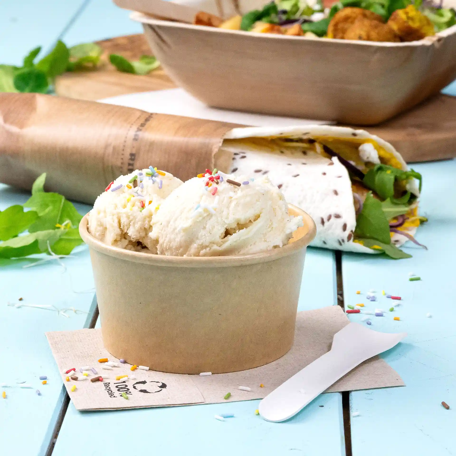 Brown paper ice cream cup with two scoops of ice cream topped with colorful sprinkles, placed on a blue surface.