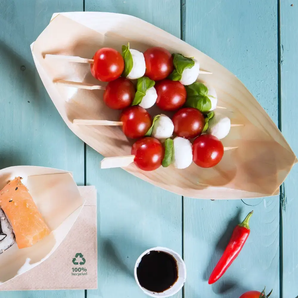 Wooden boat-shaped tray with skewers of cherry tomatoes, mozzarella balls, and basil leaves, alongside sushi and dipping sauce.