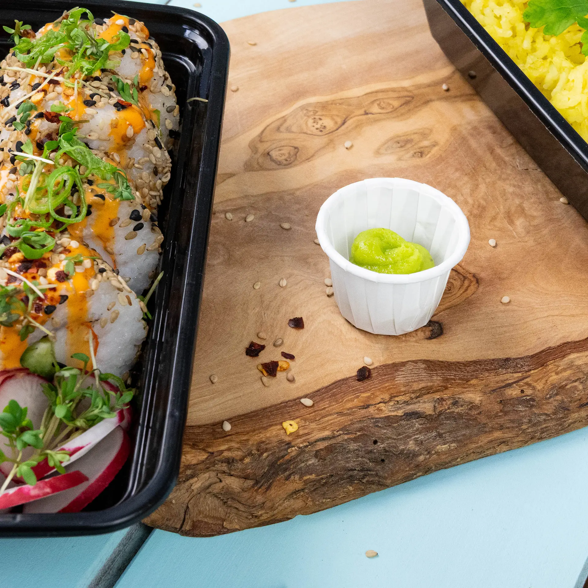 Container of sushi rolls with sesame seeds and toppings, accompanied by a small white paper cup of wasabi.