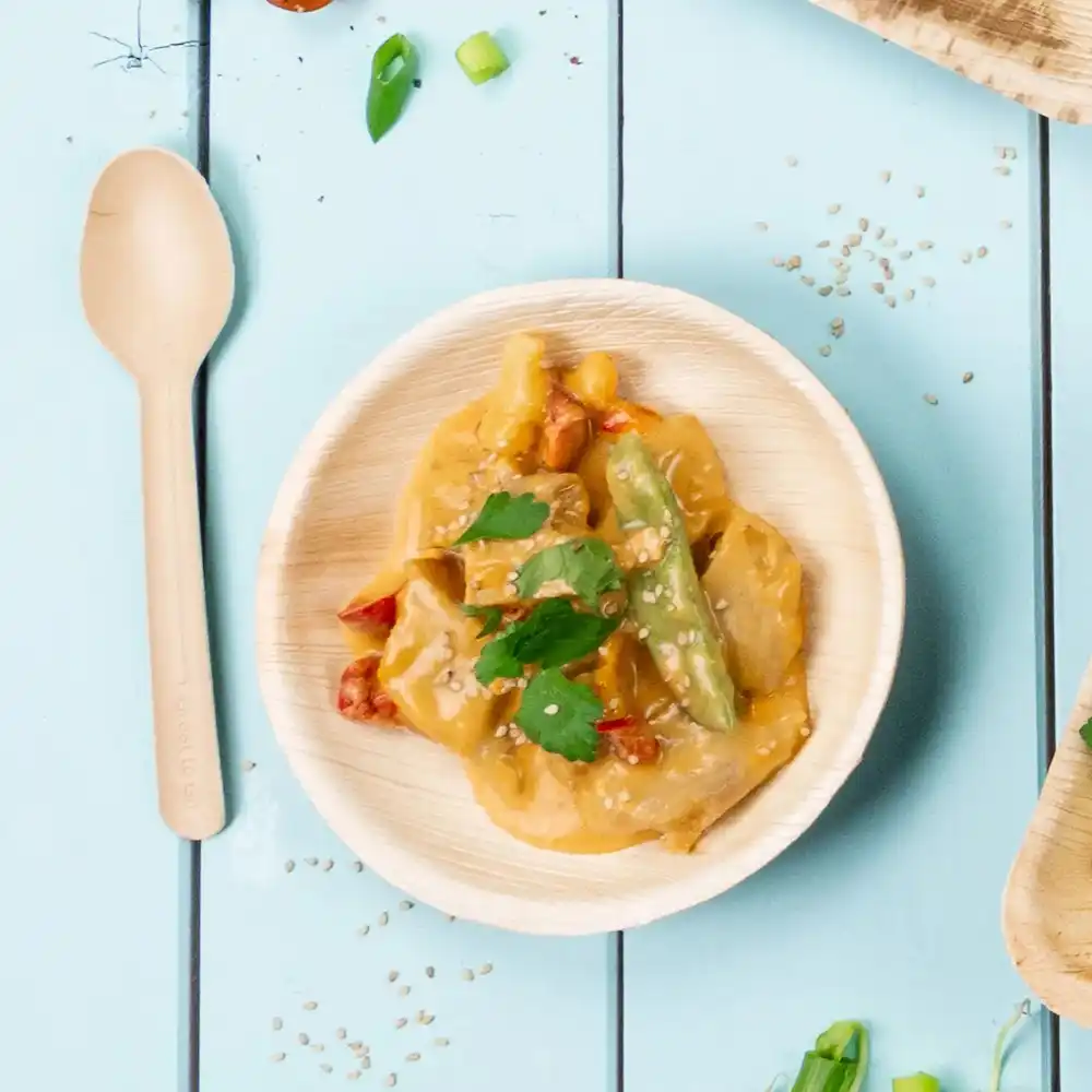 Bamboo bowl with stir-fried vegetables and sauce topped with sesame seeds and coriander, beside a wooden spoon.