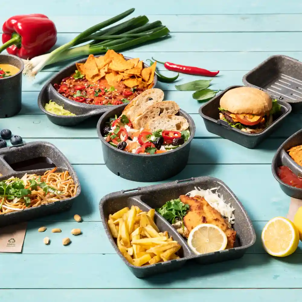 Assorted food items in black containers: nachos, salad with bread, spaghetti, fries, a burger, and additional ingredients on a wooden table.
