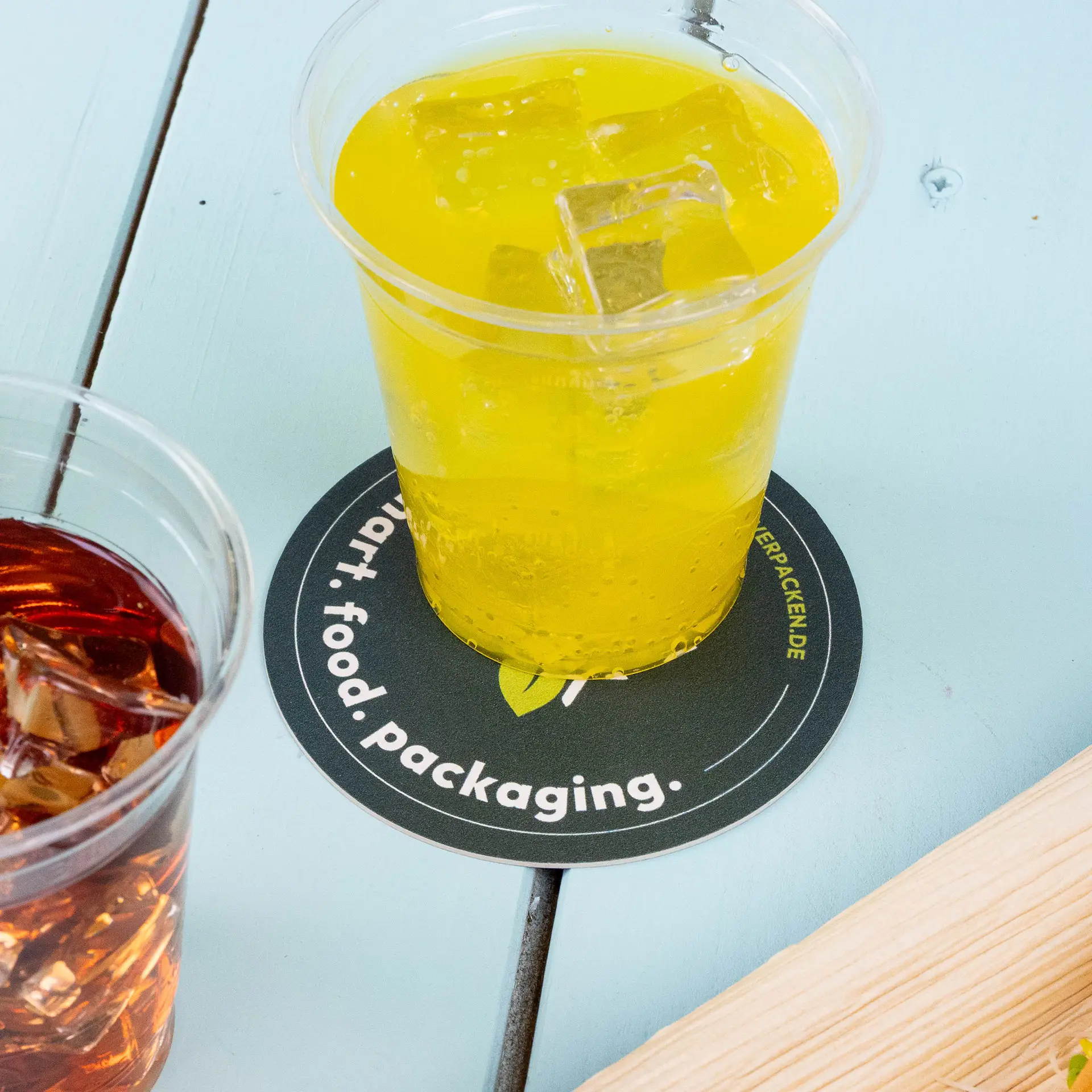 Yellow beverage in a clear cup on a round printed coasters with text, next to a cup of dark drink with ice.