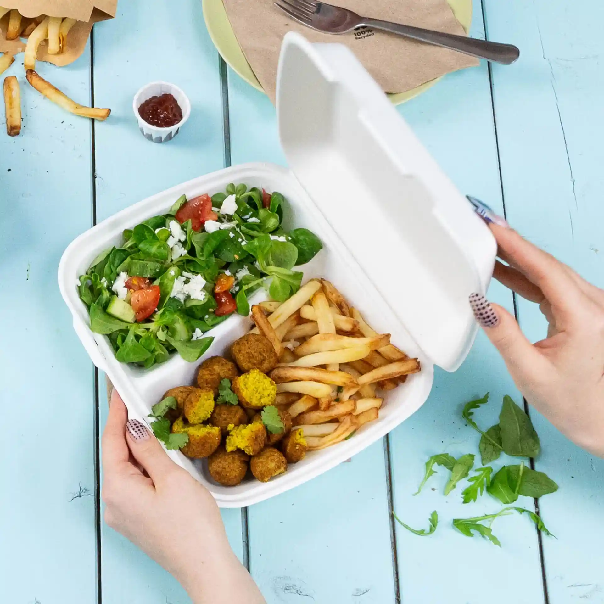 A hand holding a white hinged-lid container with two compartments, filled with salad, fries, and falafel balls.