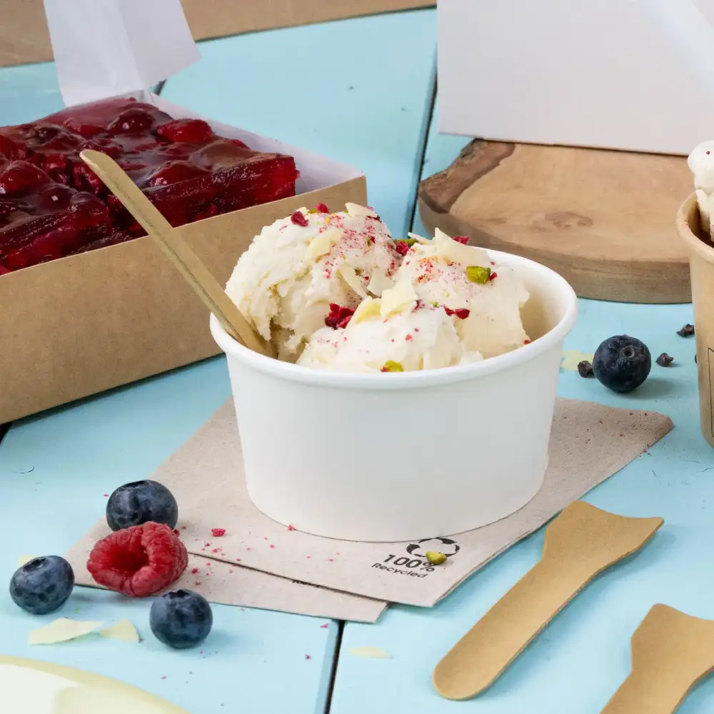 Ice cream in a white cup topped with nuts and berries beside a dessert box, with wooden spoons and fresh fruit on a blue surface.