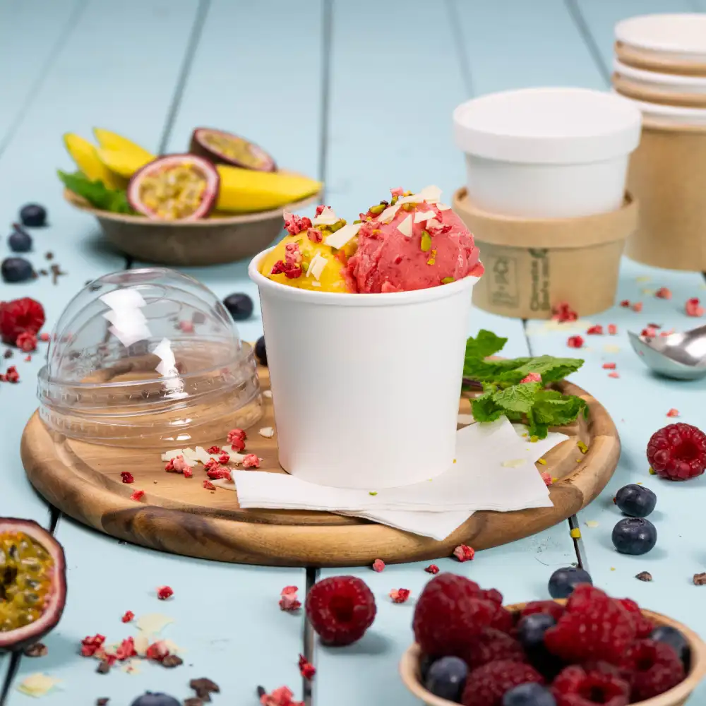 White cup filled with mango and raspberry ice cream, topped with nuts and surrounded by fresh fruit and scattered toppings.