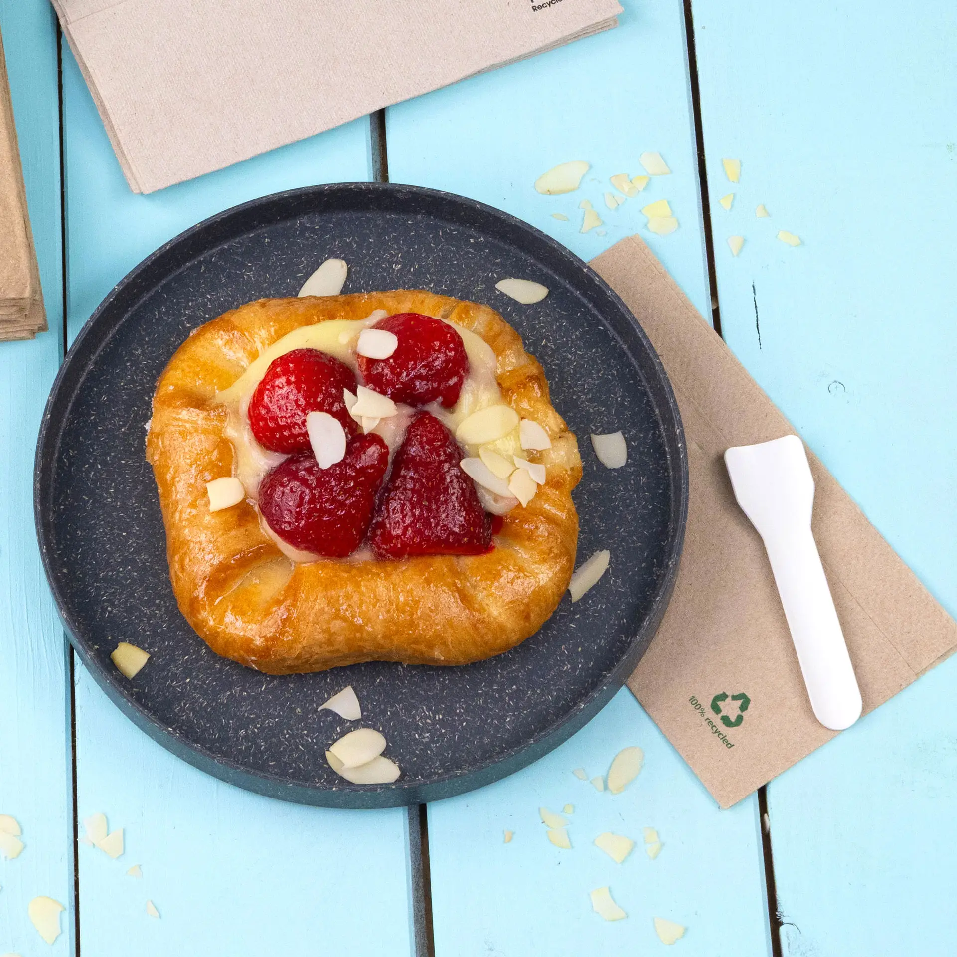 Pastry with strawberries and almond slices on a dark plate, accompanied by a white plastic utensil and unbleached napkin.