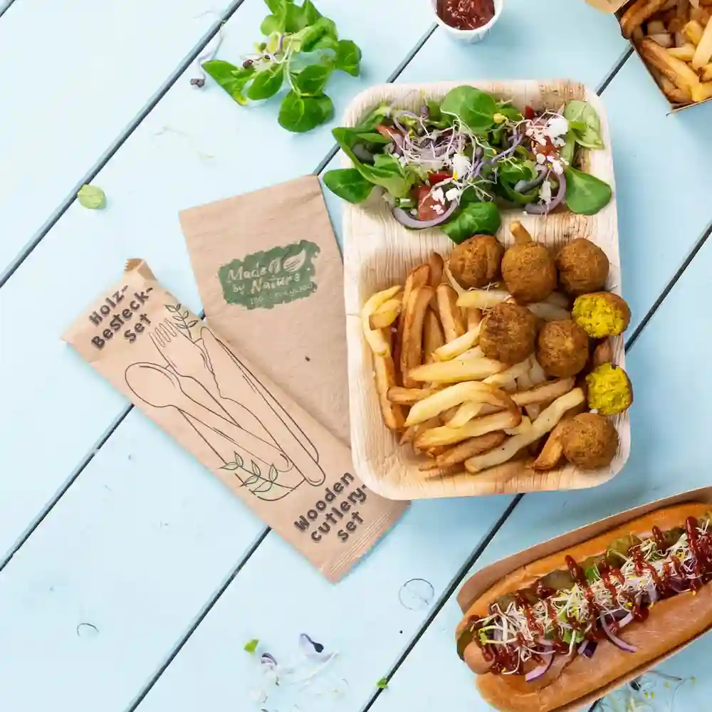 Wooden cutlery set next to a food platter with salad, fries, falafel balls, and a hot dog on a blue table.