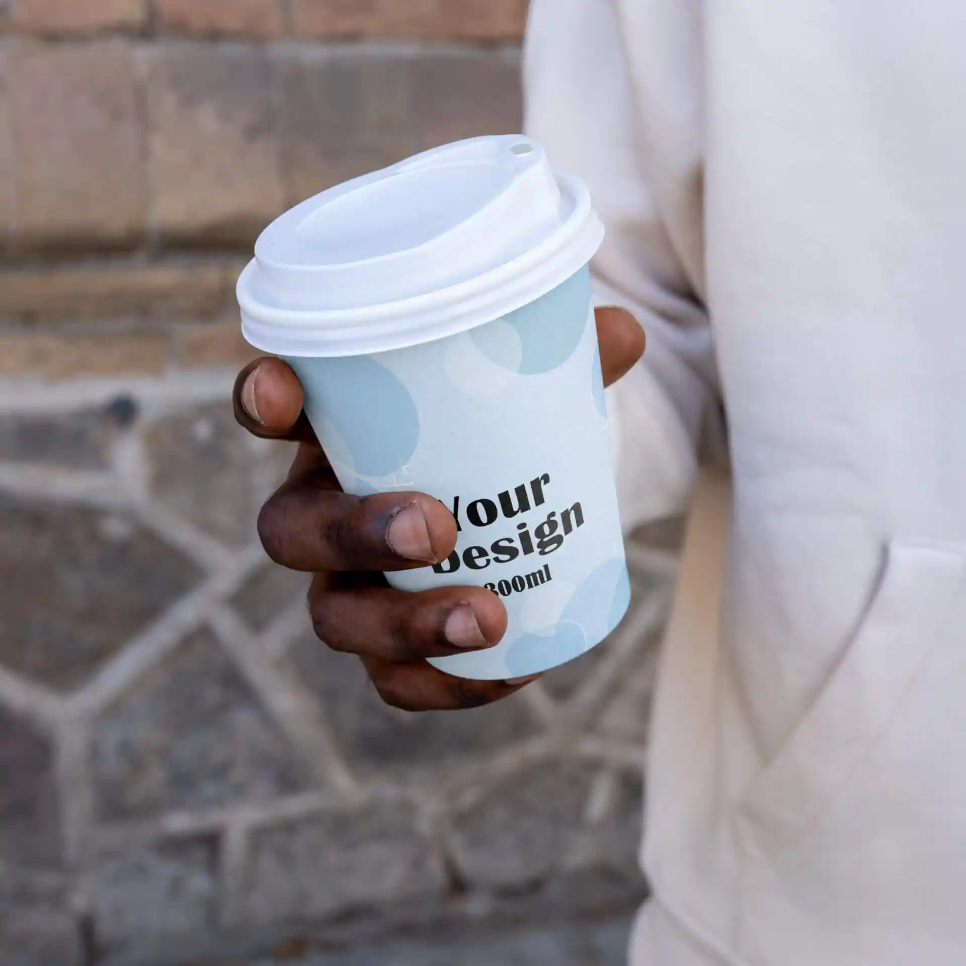 A hand holding a white paper cup with a light blue design and a white lid, featuring the text "Your design" and a volume of 300 ml.