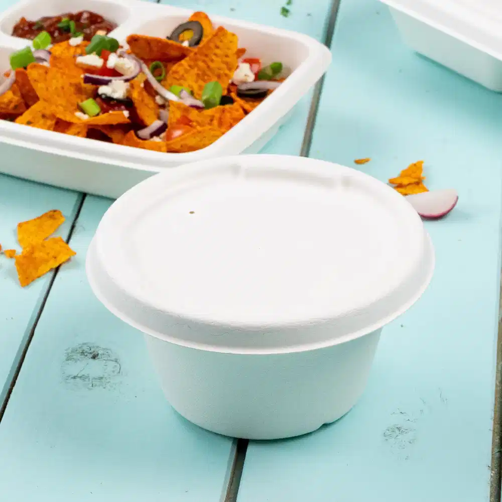 White paper soup cup on a blue tabletop with a background of nachos and a sealed food container.