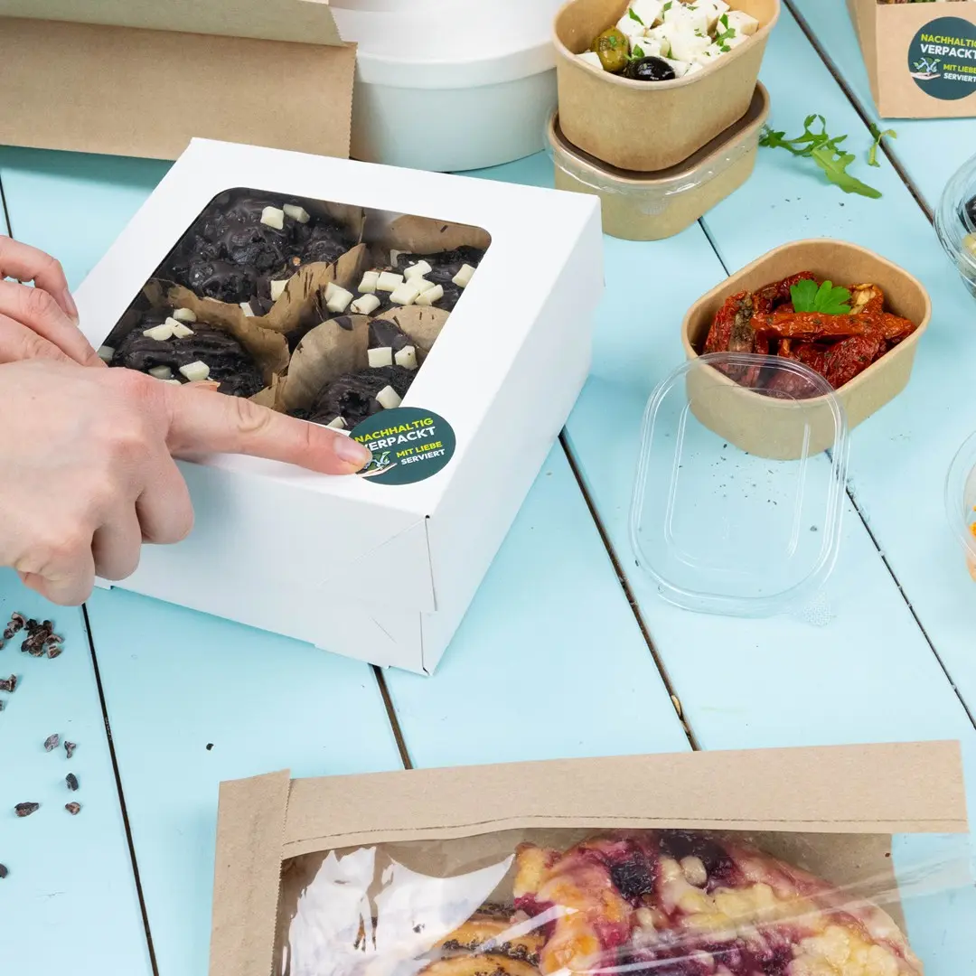 White box containing baked goods with a clear lid, surrounded by various food containers on a blue table.
