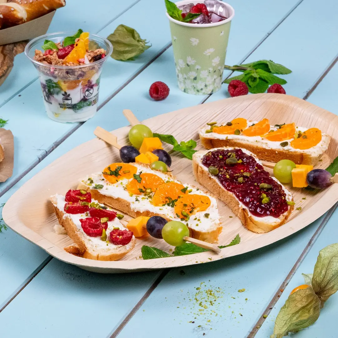 Platter with various finger foods including bread topped with cream cheese, fruits, and a fruit skewer, alongside a yogurt parfait.