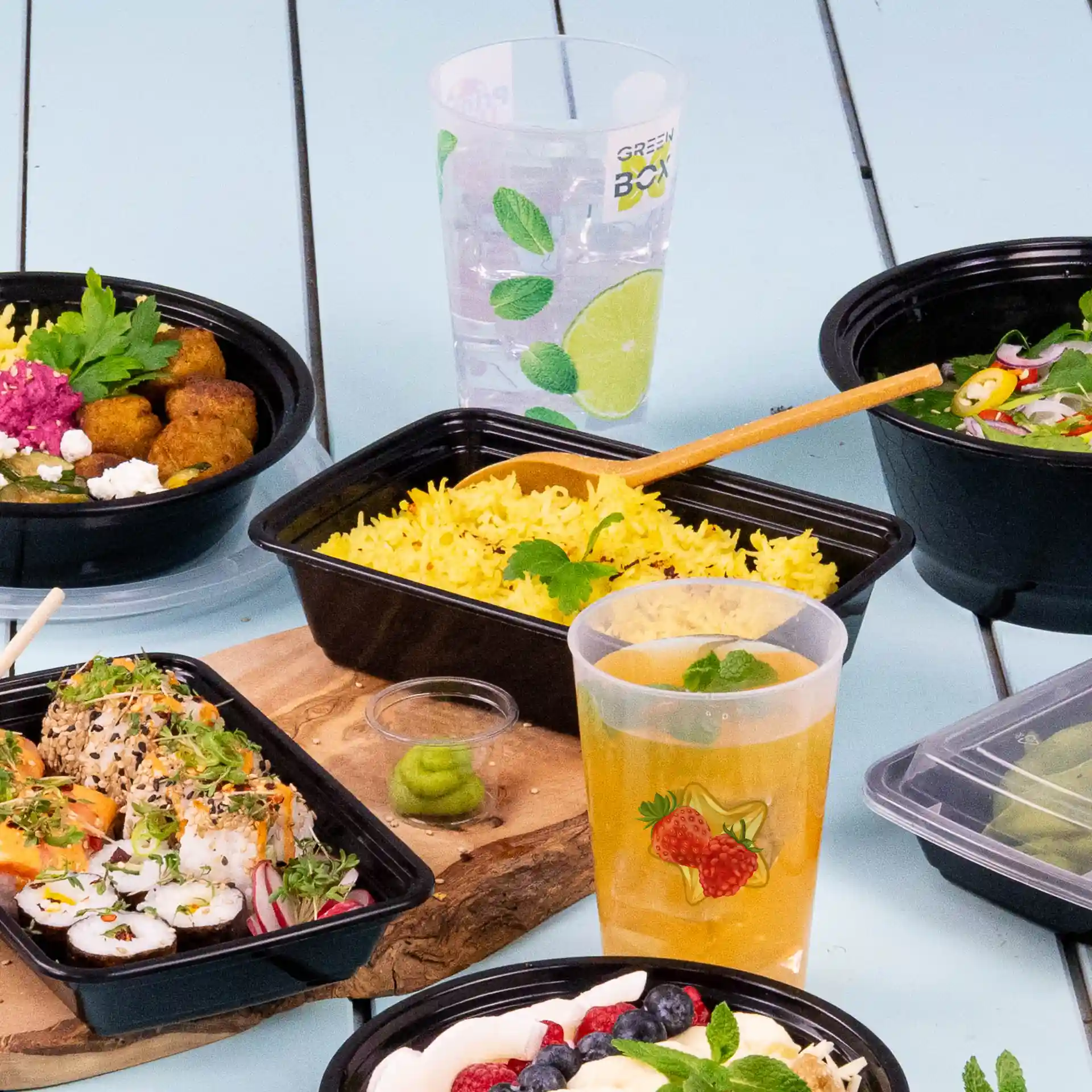 Clear reusable cup with logo, filled with a drink, surrounded by various food containers on a blue surface.