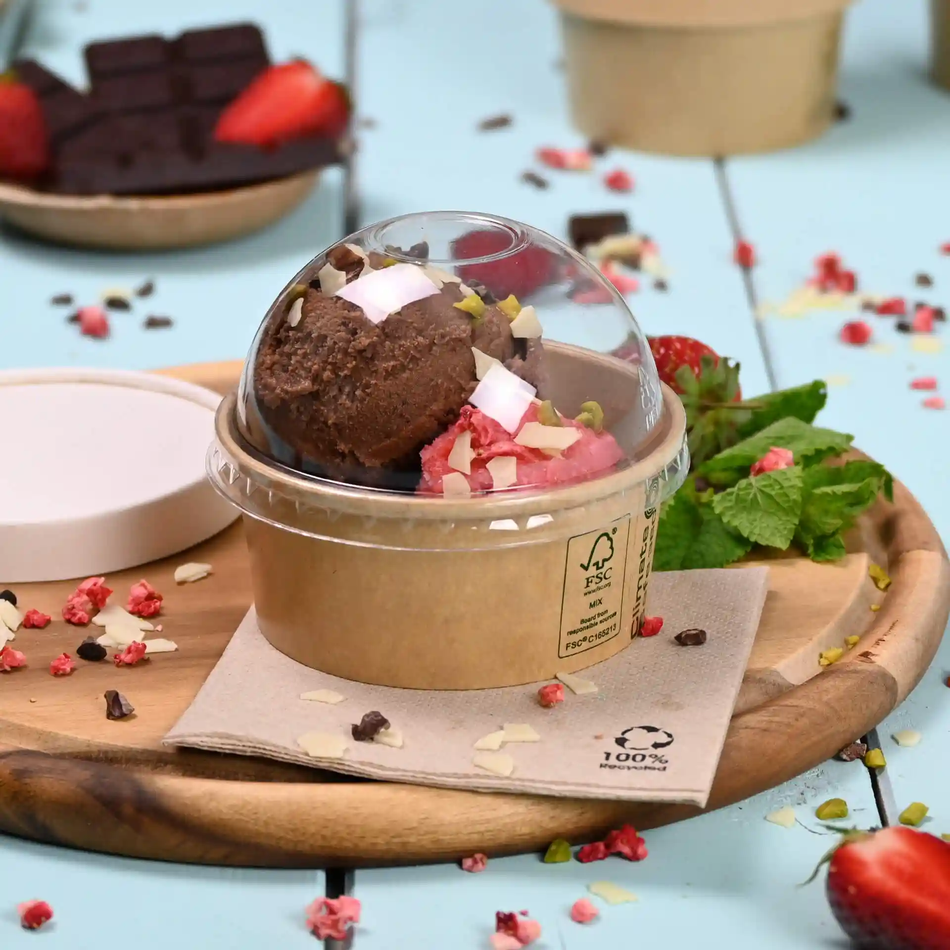 Container with two scoops of ice cream, topped with nuts and coconut, on a wooden board with mint and strawberries.