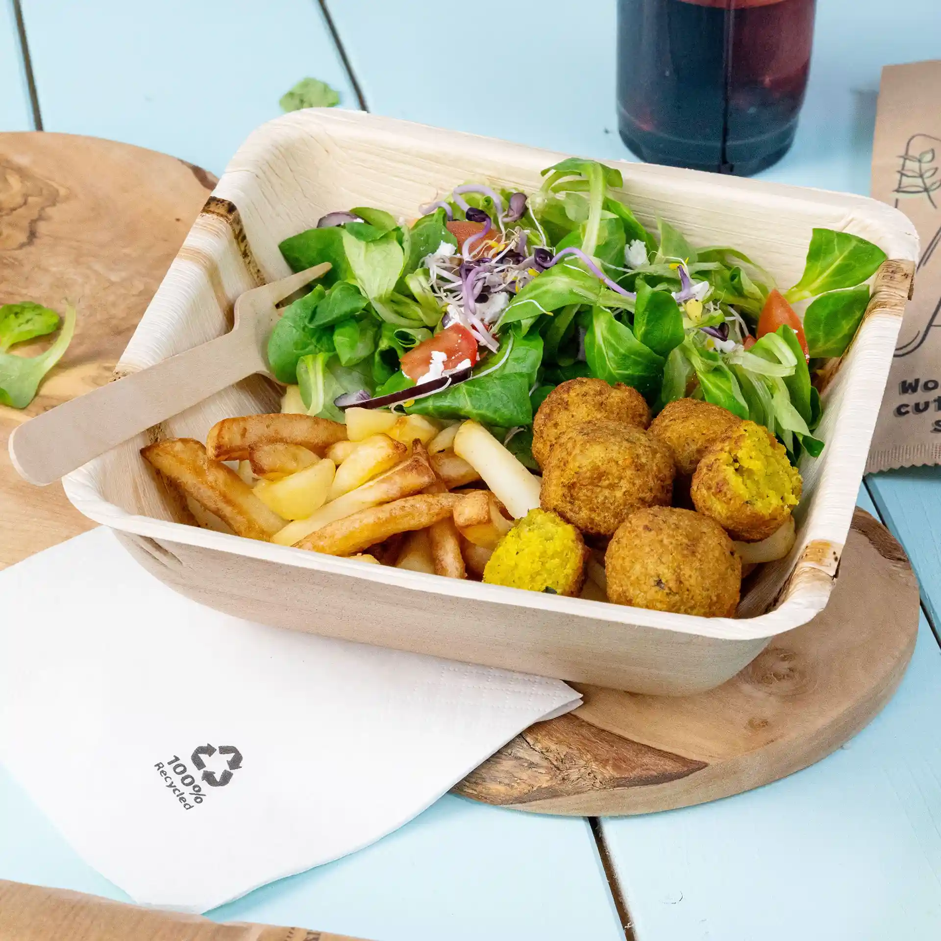 Deep square palm leaf bowl containing salad, fries, and falafel balls, with a wooden fork and a napkin beside it.