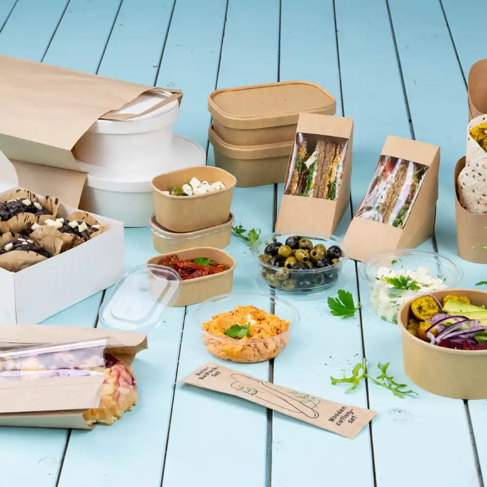 An assortment of food containers includes paper boxes, bowls, and clear plastic cups with various dishes and snacks.