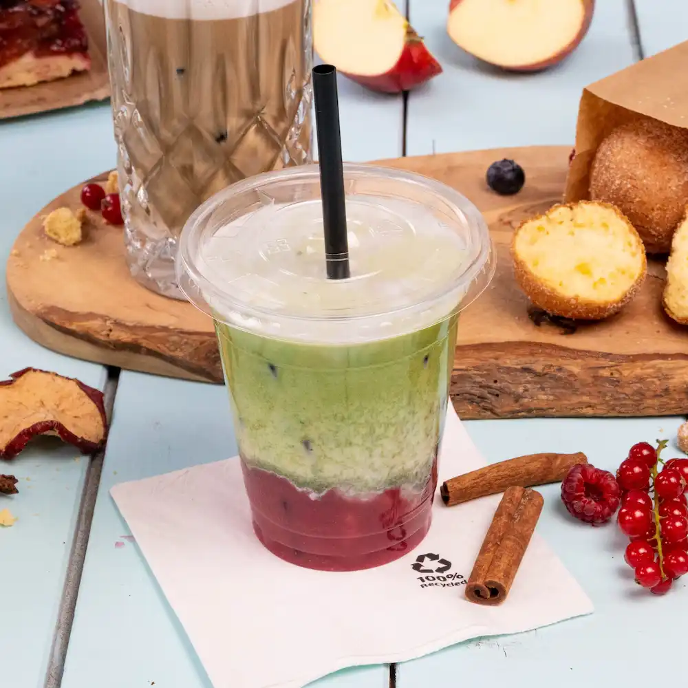 Iced layered drink in a transparent cup with a straw, surrounded by snacks and a napkin on a light blue surface.