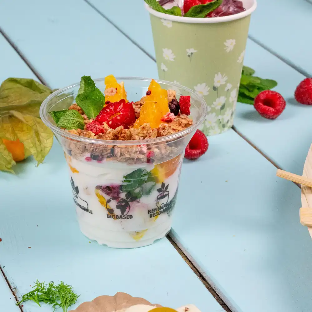 Clear plastic dessert cup filled with yogurt, fruits, and granola, placed on a blue surface with mint leaves nearby.