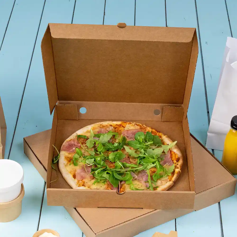 Pizza in a cardboard takeout box topped with ham and mixed greens on a blue wooden surface.