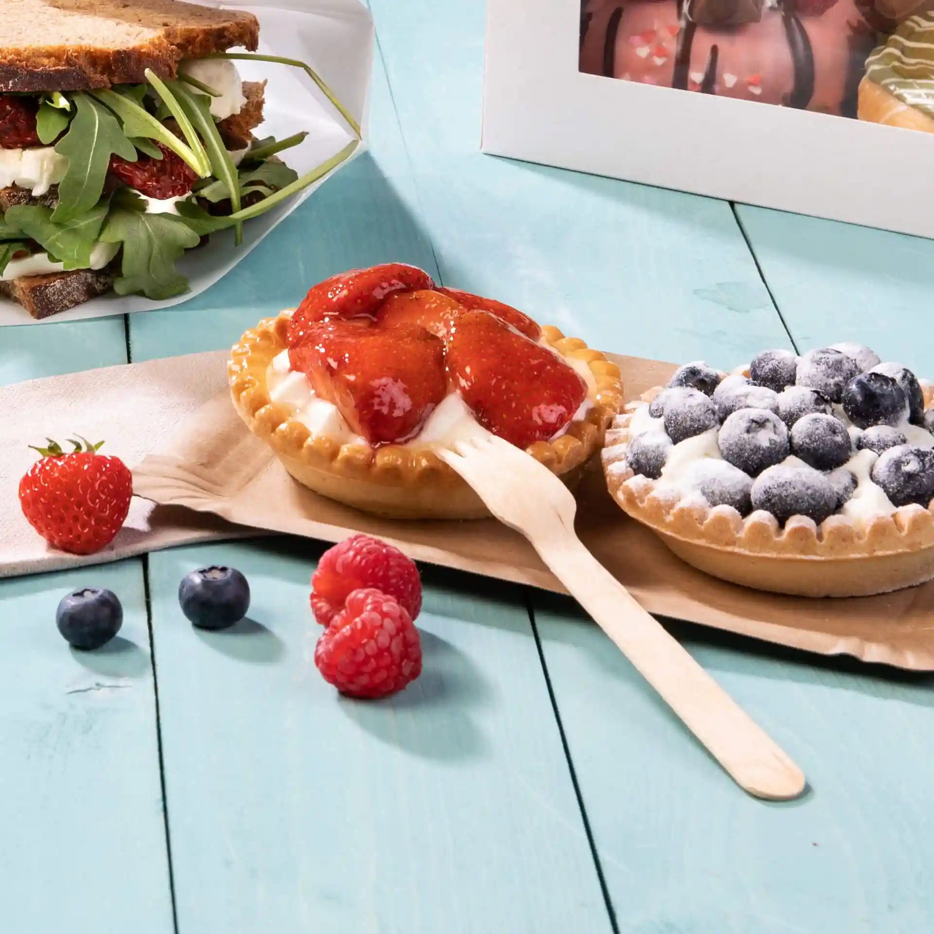 Image of two dessert tarts with strawberries and blueberries, a wooden fork, and fresh berries on a blue wooden surface.